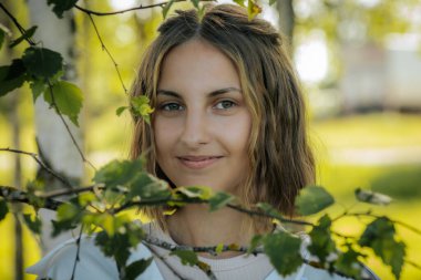 Yeşil gözlü, doğal ve sakin bir yüz ifadesi olan bir kadının yakın çekimi. Arka plan bulanık yeşillik gösteriyor, yüz hatlarına odaklanmasını sağlıyor..