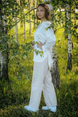 Beyaz gömlekli bir kadın yeşil yapraklarla çevrili huş ağaçları arasında güven içinde poz veriyor. Güneş ışığı, orman ortamında sıcak ve doğal bir ortam yaratır..