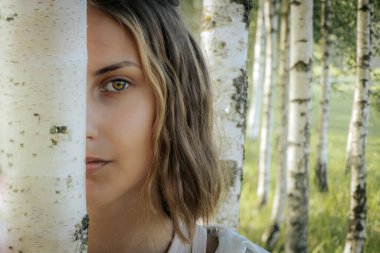 Yeşil gözlü bir kadın ormanda bir huş ağacının arkasından esrarengiz ve doğal bir manzara yaratıyor. Arka planda bulanık bir orman var, derinlik ve dinginlik katıyor..