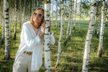 Güneş gözlüğü takan gülümseyen bir kadın güneşli bir ormanda bir huş ağacına yaslanır ve huzurlu ve neşeli bir anı yakalar..