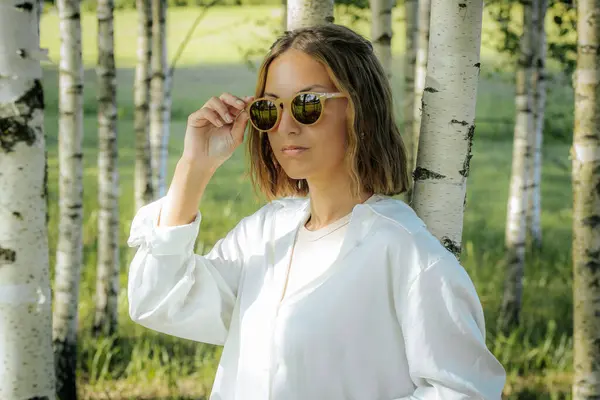 stock image A stylish woman in a white shirt adjusts her sunglasses while standing in a birch tree forest. The sunlight and natural background reflect off her glasses, creating a cool and fashionable look.