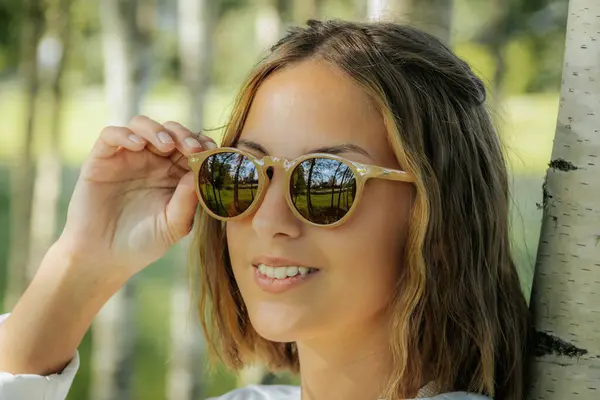 Stock image A stylish woman in a white shirt adjusts her sunglasses while standing in a birch tree forest. The sunlight and natural background reflect off her glasses, creating a cool and fashionable look.