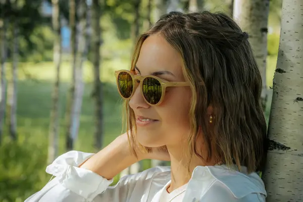 stock image A stylish woman in a white shirt adjusts her sunglasses while standing in a birch tree forest. The sunlight and natural background reflect off her glasses, creating a cool and fashionable look.