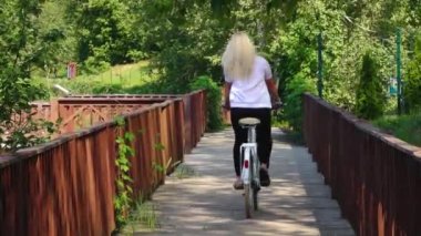 Uzun sarı saçlı bir kadın tahta bir tahta kaldırımda bisiklet sürüyor. Sahne, güneş ışığı yolu ve doğal çevresiyle özgürlük ve macera hissini yakalıyor.
