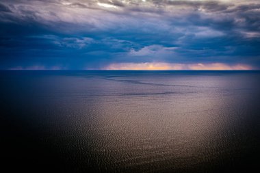 Gün batımında sakin bir denizin üzerinde, çarpıcı sarı ve turuncu bir ufka sahip, karanlık ve fırtınalı bulutlarla dramatik bir gökyüzü. Görüntü, sakin su ve çalkantılı gökyüzü arasındaki zıtlığı yakalar..