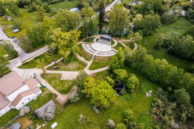 Dikli Müzik Sahnesi 'nin yemyeşil bir parktaki hava görüntüsü. Amfitiyatro tarzı sahne iyi korunan yollar, ağaçlar ve açık çimenlik alanlarla çevrilidir. Konut binaları ve park yeri 