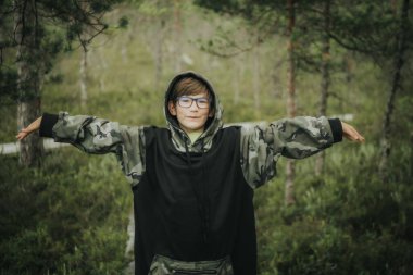 Gözlüklü ve mor kenarlı siyah kova şapkalı bir çocuk, kamuflaj kapüşonlusu giymiş, doğal bir ortamda tahta bir bankta dinleniyor..