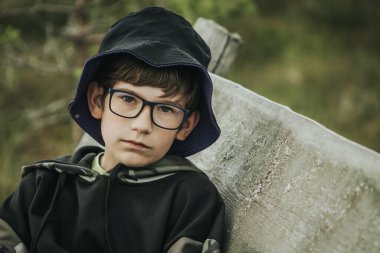Gözlüklü ve mor kenarlı siyah kova şapkalı bir çocuk, kamuflaj kapüşonlusu giymiş, doğal bir ortamda tahta bir bankta dinleniyor..