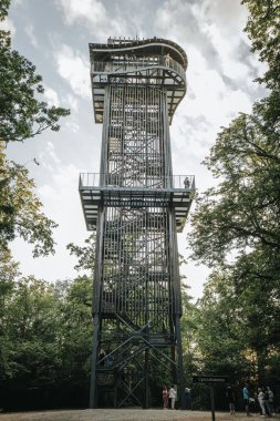 Letonya 'daki Zilakalnas gözetleme kulesinin, açık bir gökyüzünün altında yemyeşil ağaçlarla çevrili çarpıcı manzarası. Kulenin çelik yapısı belirgin bir şekilde...
