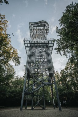Letonya 'daki Zilakalnas gözetleme kulesinin, açık bir gökyüzünün altında yemyeşil ağaçlarla çevrili çarpıcı manzarası. Kulenin çelik yapısı belirgin bir şekilde...