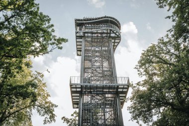 Letonya 'daki Zilakalnas gözetleme kulesinin, açık bir gökyüzünün altında yemyeşil ağaçlarla çevrili çarpıcı manzarası. Kulenin çelik yapısı belirgin bir şekilde...