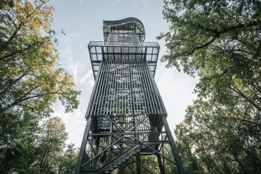 Letonya 'daki Zilakalnas gözetleme kulesinin, açık bir gökyüzünün altında yemyeşil ağaçlarla çevrili çarpıcı manzarası. Kulenin çelik yapısı belirgin bir şekilde...
