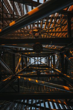 Ilık güneş ışığı bir yapının metal iskeletinde parıldar, kirişlerin üzerine altın renkler ve gölgeler bırakır. Yeşilliğin yemyeşil manzarası boşluklardan görülebilir. Boşluğu kopyala.