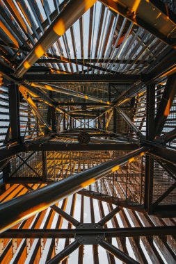 Ilık güneş ışığı bir yapının metal iskeletinde parıldar, kirişlerin üzerine altın renkler ve gölgeler bırakır. Yeşilliğin yemyeşil manzarası boşluklardan görülebilir. Boşluğu kopyala.