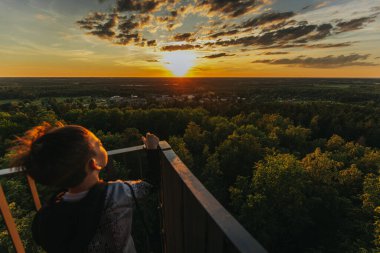 Genç bir çocuk, altın ışıkla yıkanmış manzarayla, yüksek bir bakış açısından canlı bir günbatımına bakıyor. Huzurlu an, doğanın güzelliğini ve gençliğin mucizesini yakalar..