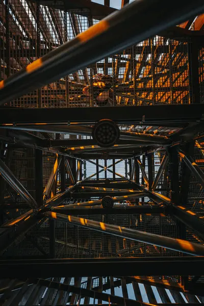 Ilık güneş ışığı bir yapının metal iskeletinde parıldar, kirişlerin üzerine altın renkler ve gölgeler bırakır. Yeşilliğin yemyeşil manzarası boşluklardan görülebilir. Boşluğu kopyala.