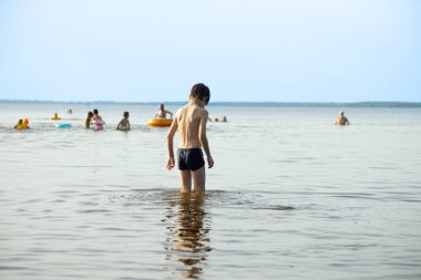 Bir çocuk sahilde sığ sularda yürüyor, etrafındaki insanlar suyun tadını çıkarıyorlar. Görüntünün sessiz, eski bir tonu var, nostaljik bir his katıyor..
