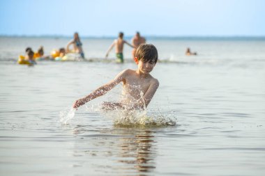 Genç bir çocuk sevinçle iki kolu kumsalda yükselirken su sıçratır ve etrafında damlacıklar oluşturur. Arka planda güneşli günün tadını çıkaran birkaç kişi var..