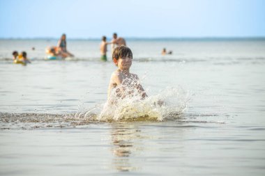 Genç bir çocuk sevinçle iki kolu kumsalda yükselirken su sıçratır ve etrafında damlacıklar oluşturur. Arka planda güneşli günün tadını çıkaran birkaç kişi var..