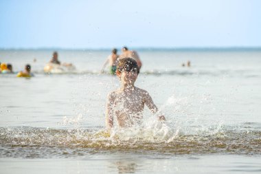 Genç bir çocuk sevinçle iki kolu kumsalda yükselirken su sıçratır ve etrafında damlacıklar oluşturur. Arka planda güneşli günün tadını çıkaran birkaç kişi var..