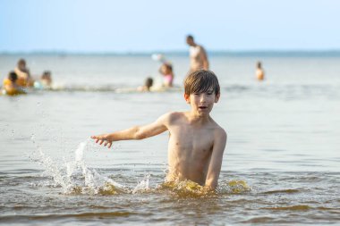 Genç bir çocuk sevinçle iki kolu kumsalda yükselirken su sıçratır ve etrafında damlacıklar oluşturur. Arka planda güneşli günün tadını çıkaran birkaç kişi var..