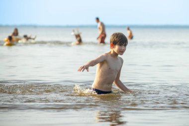 Genç bir çocuk sevinçle iki kolu kumsalda yükselirken su sıçratır ve etrafında damlacıklar oluşturur. Arka planda güneşli günün tadını çıkaran birkaç kişi var..