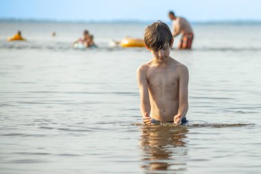 Genç bir çocuk sevinçle iki kolu kumsalda yükselirken su sıçratır ve etrafında damlacıklar oluşturur. Arka planda güneşli günün tadını çıkaran birkaç kişi var..