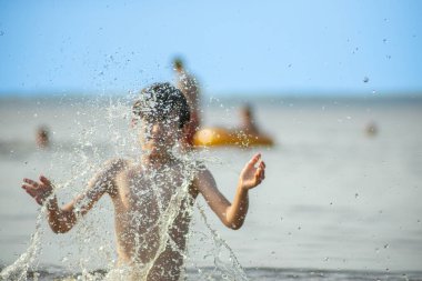 Genç bir çocuk sevinçle iki kolu kumsalda yükselirken su sıçratır ve etrafında damlacıklar oluşturur. Arka planda güneşli günün tadını çıkaran birkaç kişi var..
