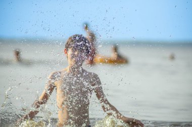 Genç bir çocuk sevinçle iki kolu kumsalda yükselirken su sıçratır ve etrafında damlacıklar oluşturur. Arka planda güneşli günün tadını çıkaran birkaç kişi var..