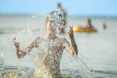 Genç bir çocuk sevinçle iki kolu kumsalda yükselirken su sıçratır ve etrafında damlacıklar oluşturur. Arka planda güneşli günün tadını çıkaran birkaç kişi var..