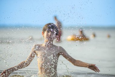 Genç bir çocuk sevinçle iki kolu kumsalda yükselirken su sıçratır ve etrafında damlacıklar oluşturur. Arka planda güneşli günün tadını çıkaran birkaç kişi var..