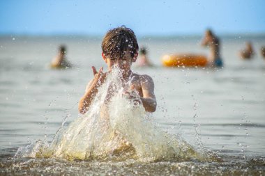 Genç bir çocuk sevinçle iki kolu kumsalda yükselirken su sıçratır ve etrafında damlacıklar oluşturur. Arka planda güneşli günün tadını çıkaran birkaç kişi var..