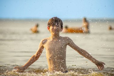 Genç bir çocuk sevinçle iki kolu kumsalda yükselirken su sıçratır ve etrafında damlacıklar oluşturur. Arka planda güneşli günün tadını çıkaran birkaç kişi var..