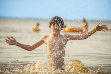 Genç bir çocuk sevinçle iki kolu kumsalda yükselirken su sıçratır ve etrafında damlacıklar oluşturur. Arka planda güneşli günün tadını çıkaran birkaç kişi var..