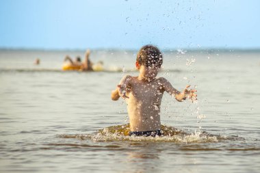 Genç bir çocuk sevinçle iki kolu kumsalda yükselirken su sıçratır ve etrafında damlacıklar oluşturur. Arka planda güneşli günün tadını çıkaran birkaç kişi var..