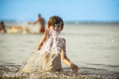 Genç bir çocuk sevinçle iki kolu kumsalda yükselirken su sıçratır ve etrafında damlacıklar oluşturur. Arka planda güneşli günün tadını çıkaran birkaç kişi var..