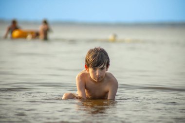Bir çocuk sahilde sığ sularda yürüyor, etrafındaki insanlar suyun tadını çıkarıyorlar. Görüntünün sessiz, eski bir tonu var, nostaljik bir his katıyor..