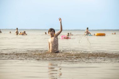 Genç bir çocuk sevinçle iki kolu kumsalda yükselirken su sıçratır ve etrafında damlacıklar oluşturur. Arka planda güneşli günün tadını çıkaran birkaç kişi var..
