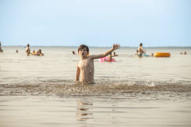 Genç bir çocuk sevinçle iki kolu kumsalda yükselirken su sıçratır ve etrafında damlacıklar oluşturur. Arka planda güneşli günün tadını çıkaran birkaç kişi var..
