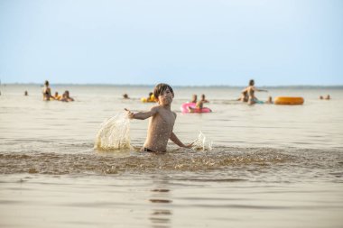 Genç bir çocuk sevinçle iki kolu kumsalda yükselirken su sıçratır ve etrafında damlacıklar oluşturur. Arka planda güneşli günün tadını çıkaran birkaç kişi var..