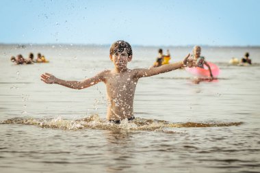Genç bir çocuk sevinçle iki kolu kumsalda yükselirken su sıçratır ve etrafında damlacıklar oluşturur. Arka planda güneşli günün tadını çıkaran birkaç kişi var..