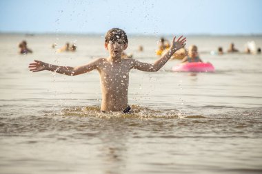 Genç bir çocuk sevinçle iki kolu kumsalda yükselirken su sıçratır ve etrafında damlacıklar oluşturur. Arka planda güneşli günün tadını çıkaran birkaç kişi var..