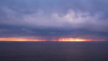 Sakin bir okyanusun üzerinde koyu, ağır bulutların altında parlak turuncu renkleri olan dramatik bir gün batımı. Ufuk, sakin su ve tehdit eden gökyüzü arasında çarpıcı bir kontrastı gösteriyor..