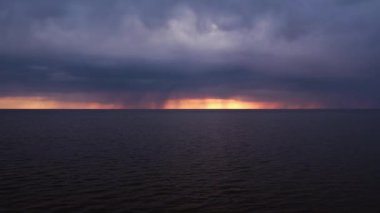 Sakin bir okyanusun üzerinde koyu, ağır bulutların altında parlak turuncu renkleri olan dramatik bir gün batımı. Ufuk, sakin su ve tehdit eden gökyüzü arasında çarpıcı bir kontrastı gösteriyor..
