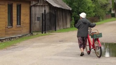 Yaşlı bir kadın, küçük bir köyde bisiklet sürüyor ve kırsal hayatın cazibesini sergiliyor. Resimli sahne geleneksel evleri ve sakin bir atmosferi yakalıyor..