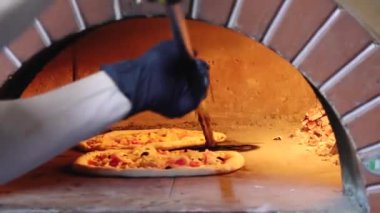 Geleneksel tuğla fırının içinde pizza pişirmenin yakın plan görüntüsü. Arka planda alevler görünüyor. Pizza çeşitli malzemelerle kaplı ve mükemmel bir şekilde pişiriliyor.