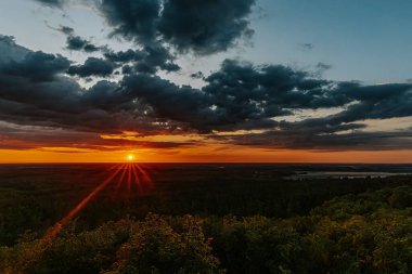 Uçsuz bucaksız orman manzarası üzerinde nefes kesici bir gün doğumu. Canlı turuncu ve sarı renkler karanlık gökyüzüne karışıyor. Dramatik bulutlar manzaraya derinlik katar. Boşluğu kopyala.