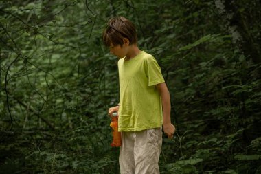 Yeşil gömlek giyen genç bir çocuk yemyeşil bir ormanda dışarıda dururken neşeyle gülümsüyor. Sahne, doğadaki masumiyet ve mutluluğu yakalar..