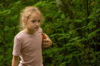 Pembe tişört giyen genç bir kız bir oyuncağı tutarak yemyeşil bir ormanı keşfediyor. Yeşillik boyunca yol alırken ifadesi bir merak ve odaklanma ifadesi..