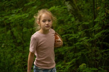 Pembe tişört giyen genç bir kız bir oyuncağı tutarak yemyeşil bir ormanı keşfediyor. Yeşillik boyunca yol alırken ifadesi bir merak ve odaklanma ifadesi..